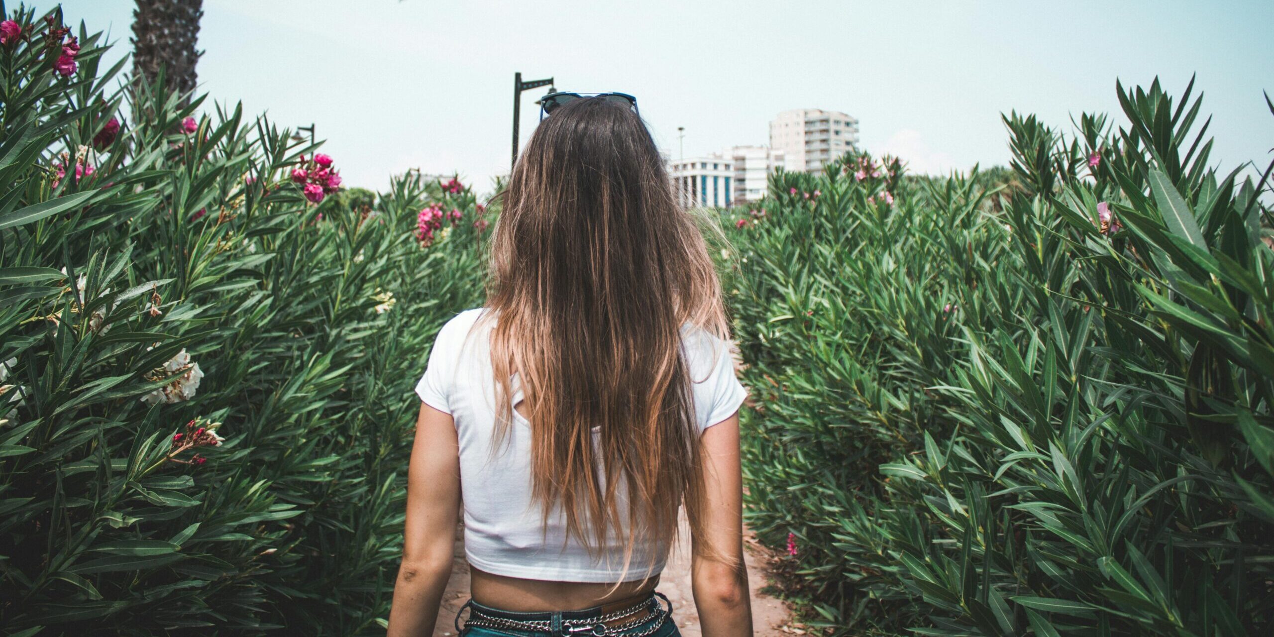 A woman in a white crop top walking through a lush garden with flowers and green plants.