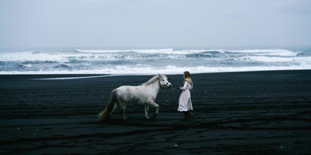 A serene scene of a woman and a horse on a black sand beach with waves in the background.