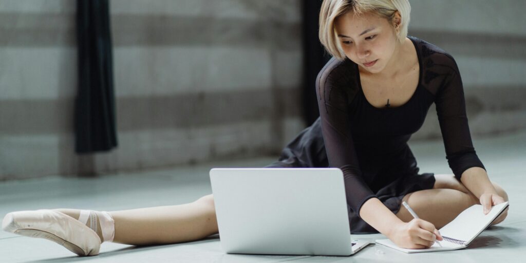 Asian ballerina studying on a laptop while taking notes in a dance studio.