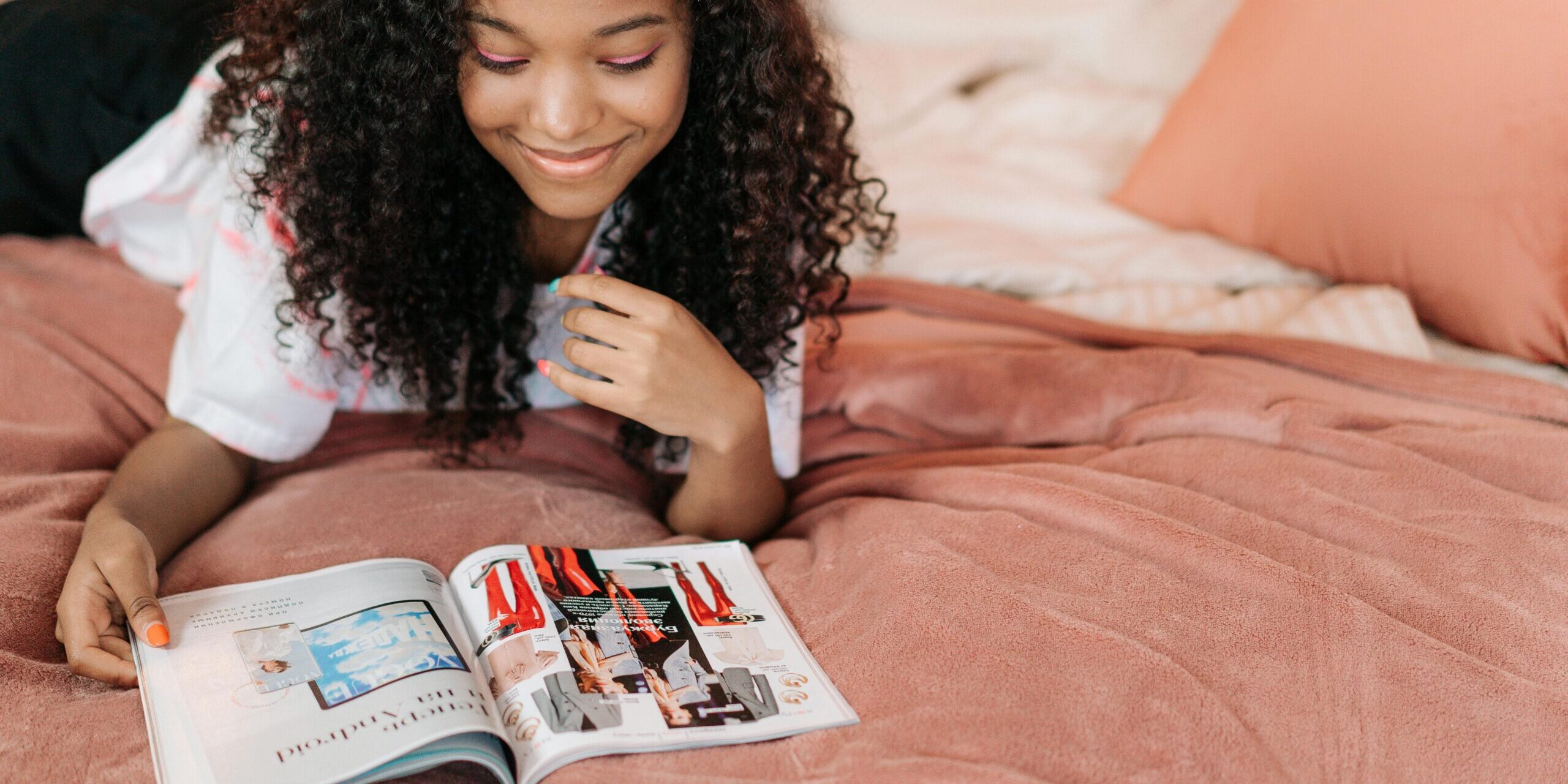 Smiling teenager with curly hair reading a magazine on a bed with peach-colored pillows.