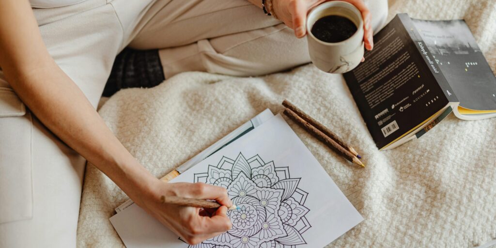 A woman enjoys a cozy moment with coloring, books, and coffee on a soft bed.