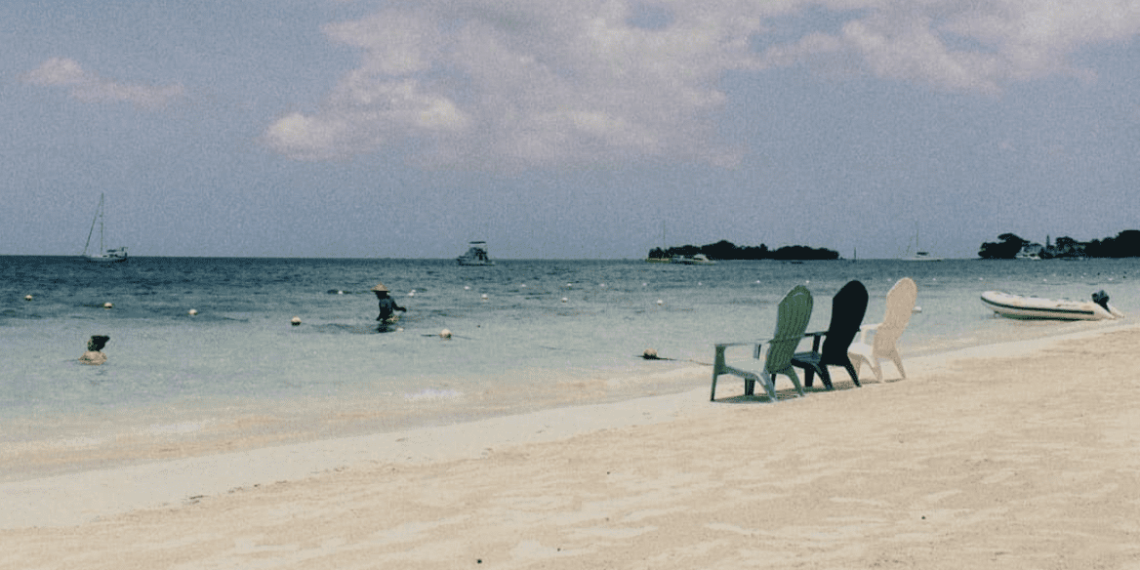 white sand beach, 3 chairs green, black, and white, boat in distance and fisherman with beige hat in water